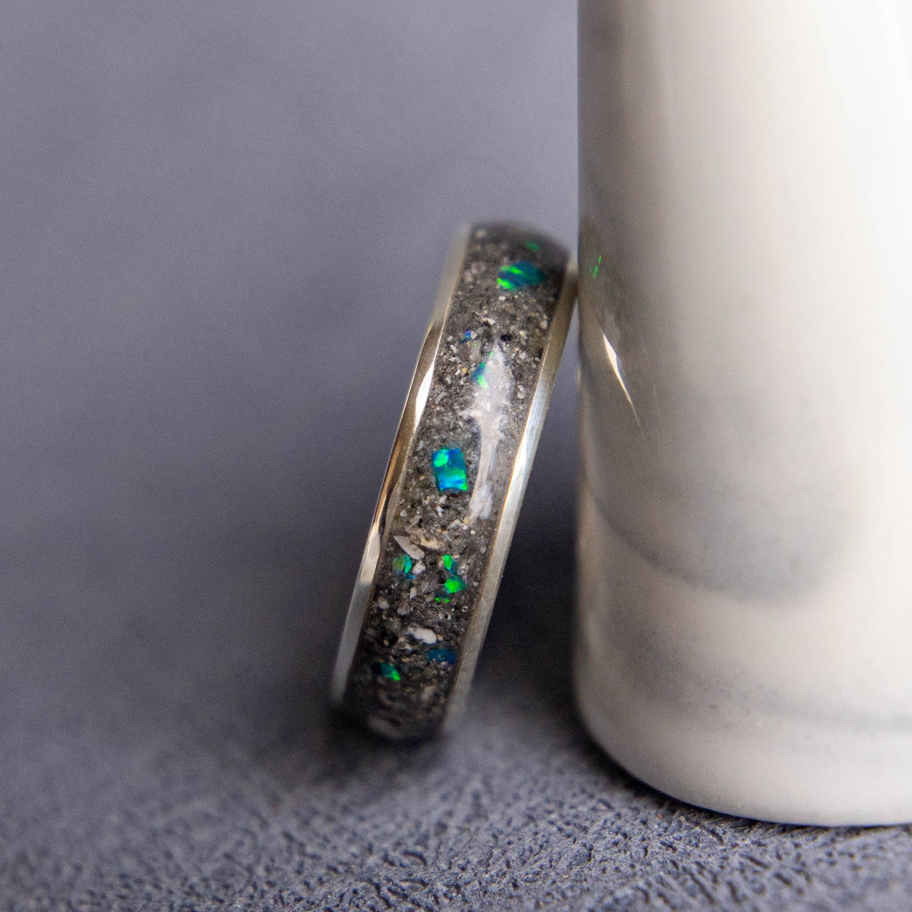 A silver-toned ring with inlaid green and blue iridescent opal-like stones, leaning against a white cylindrical object on a textured gray surface.